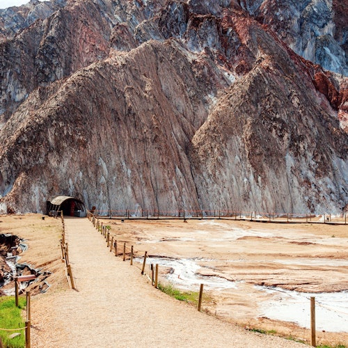 A gravel path, bordered by a simple wooden fence, leads to a tunnel entrance at the base of steep, multicolored rocky cliffs.