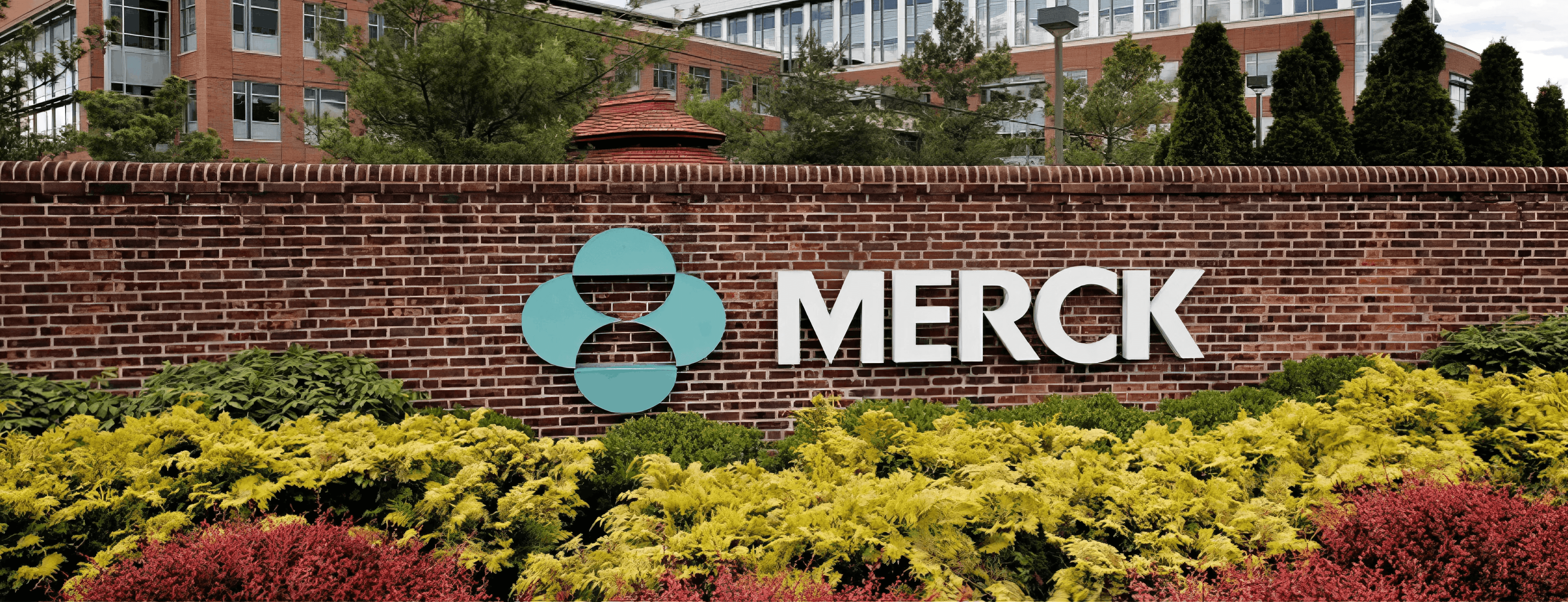 MW Interior  - Merck Unternehmenseingang mit Logo und türkisfarbenem Markenzeichen auf Ziegelsteinmauer vor mehrgeschossigem Bürogebäude" Beschreibung: Haupteingang des Merck-Standorts mit markanter Beschilderung und gepflegtem Außenbereich – ein internationaler Referenzkunde, für dessen Arbeitsumgebungen Einrichtungslösungen auf höchstem Niveau realisiert wurden.