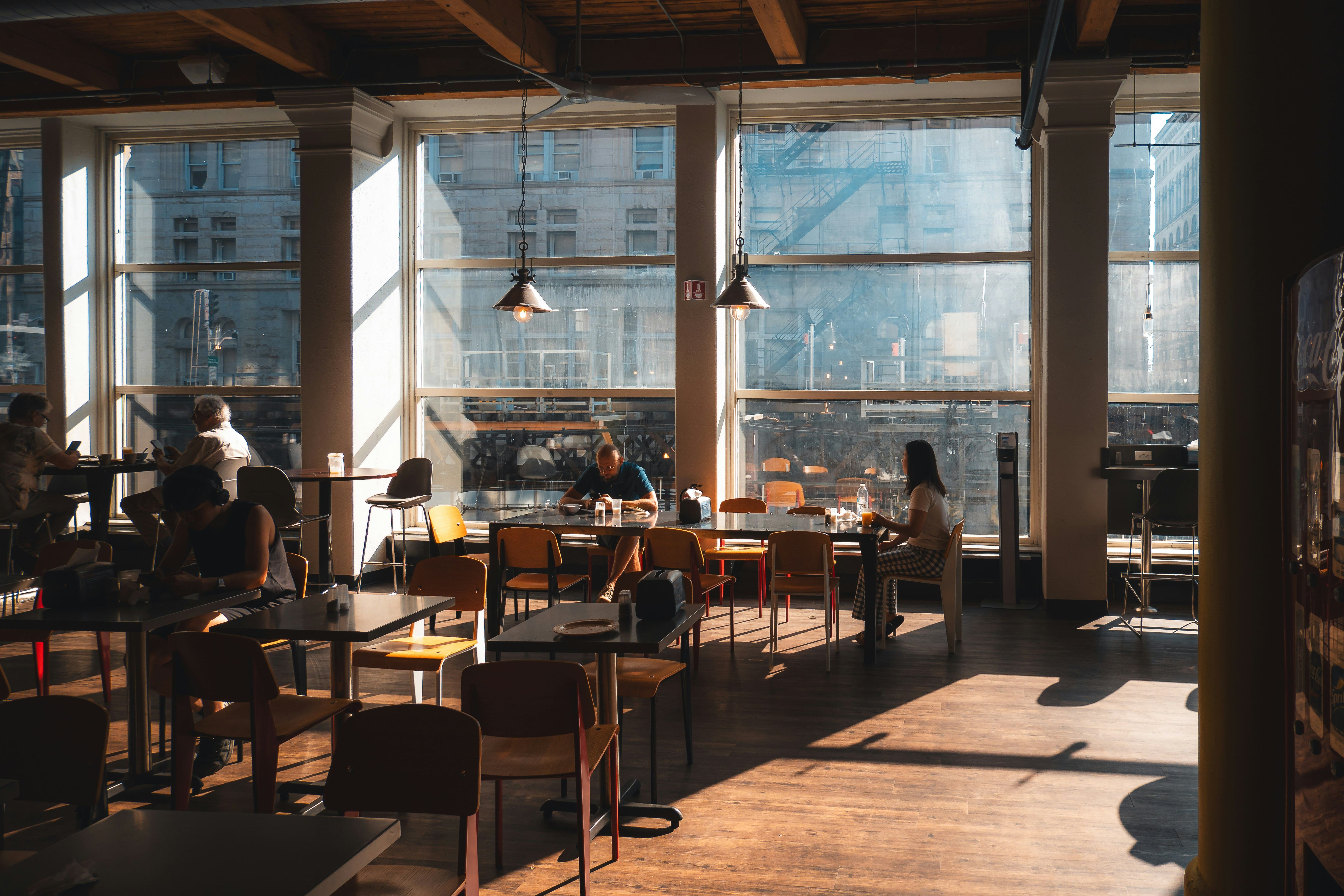 Zonnige, ruime eetzaal met grote ramen die uitkijken op een stad. Mensen zitten aan tafels, sommigen op hun telefoon. Hout...