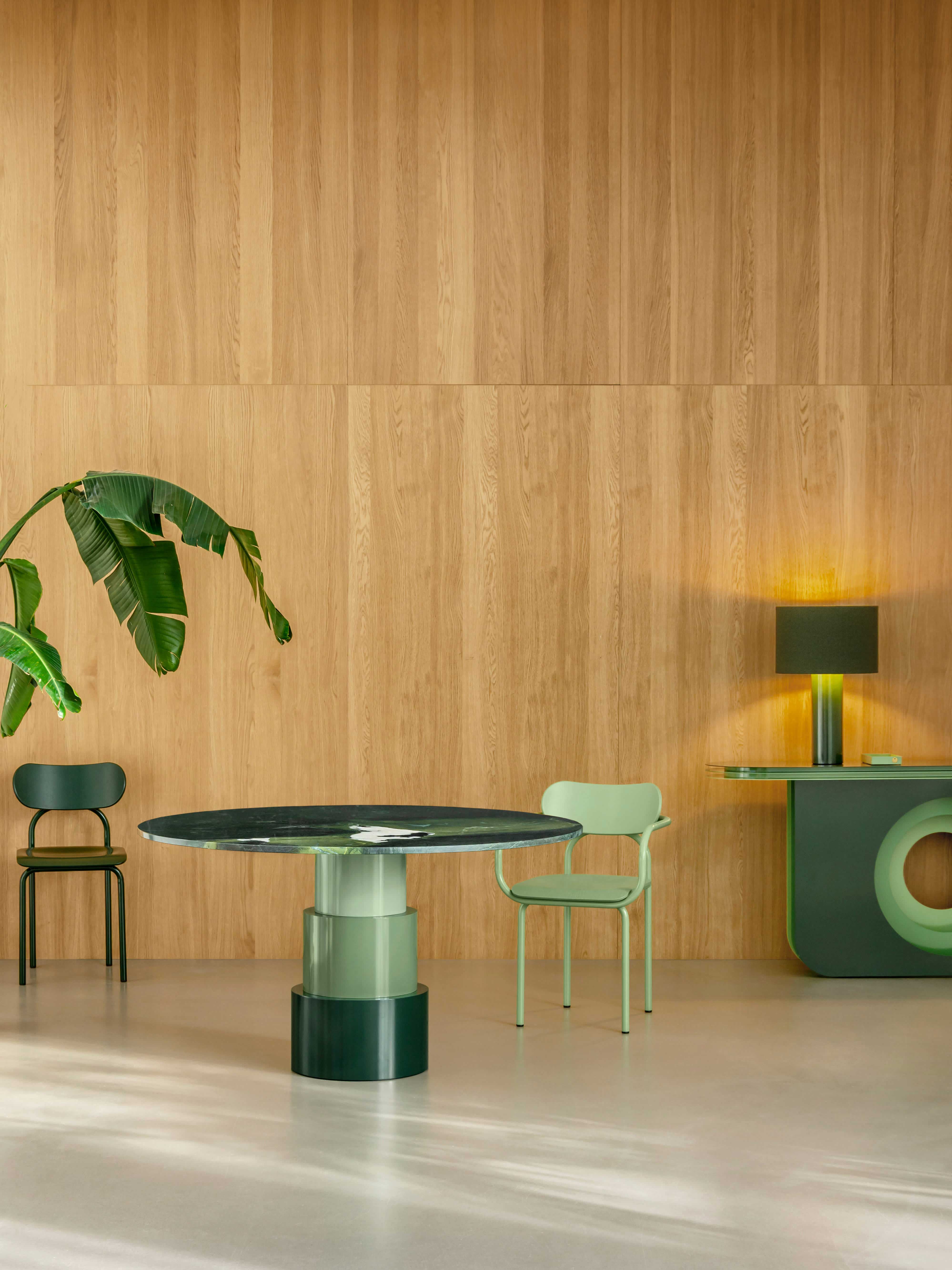 A modern dining area with a round table that has a green and white marble-like top and a light green cylindrical base. Two chairs with green seats and bases are placed around it. A tall green plant and a console table with a green table lamp are in the background, all set against a wood-paneled wall.