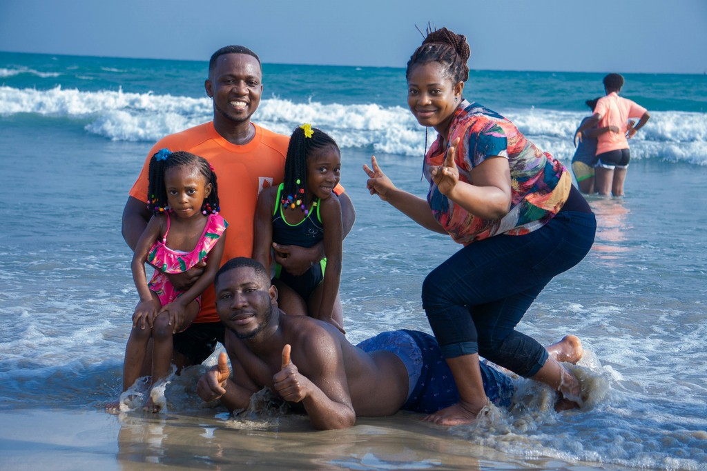 A joyful family enjoying a day at the beach, with the parents and two young girls posing in the shallow water near the shore, smiling and giving peace signs, against the backdrop of gentle waves and clear blue skies.