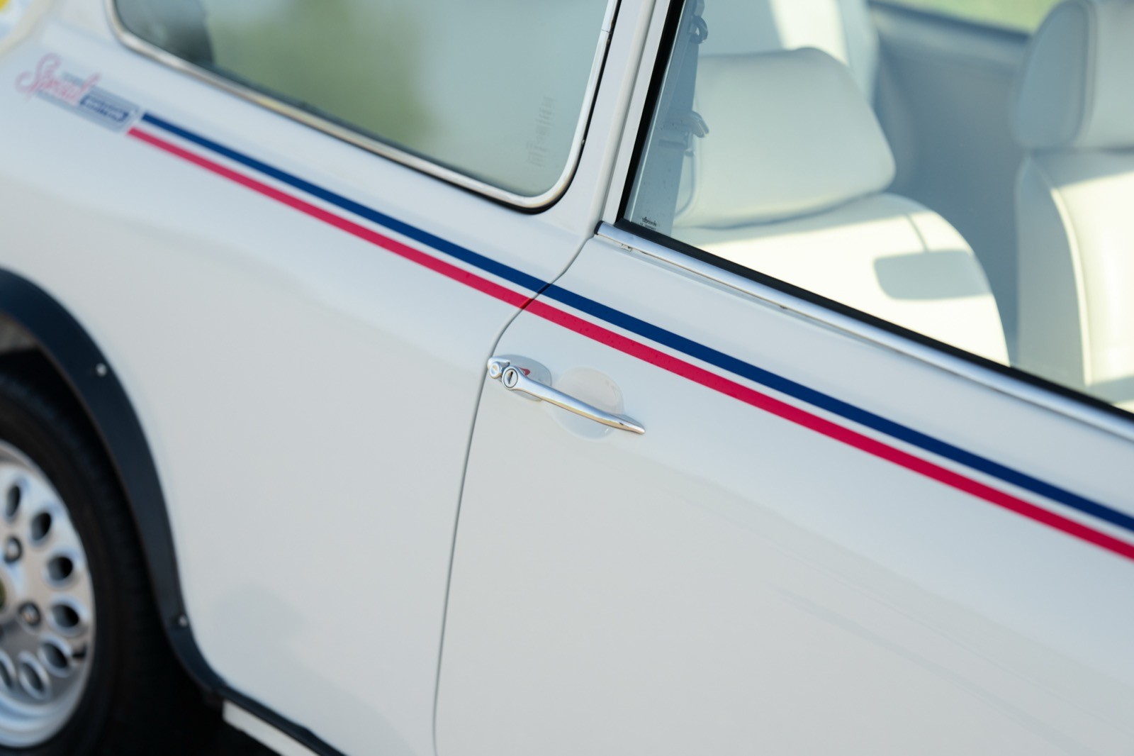 Close-up of a white car door with a red and blue stripe, showcasing the handle and part of the wheel.