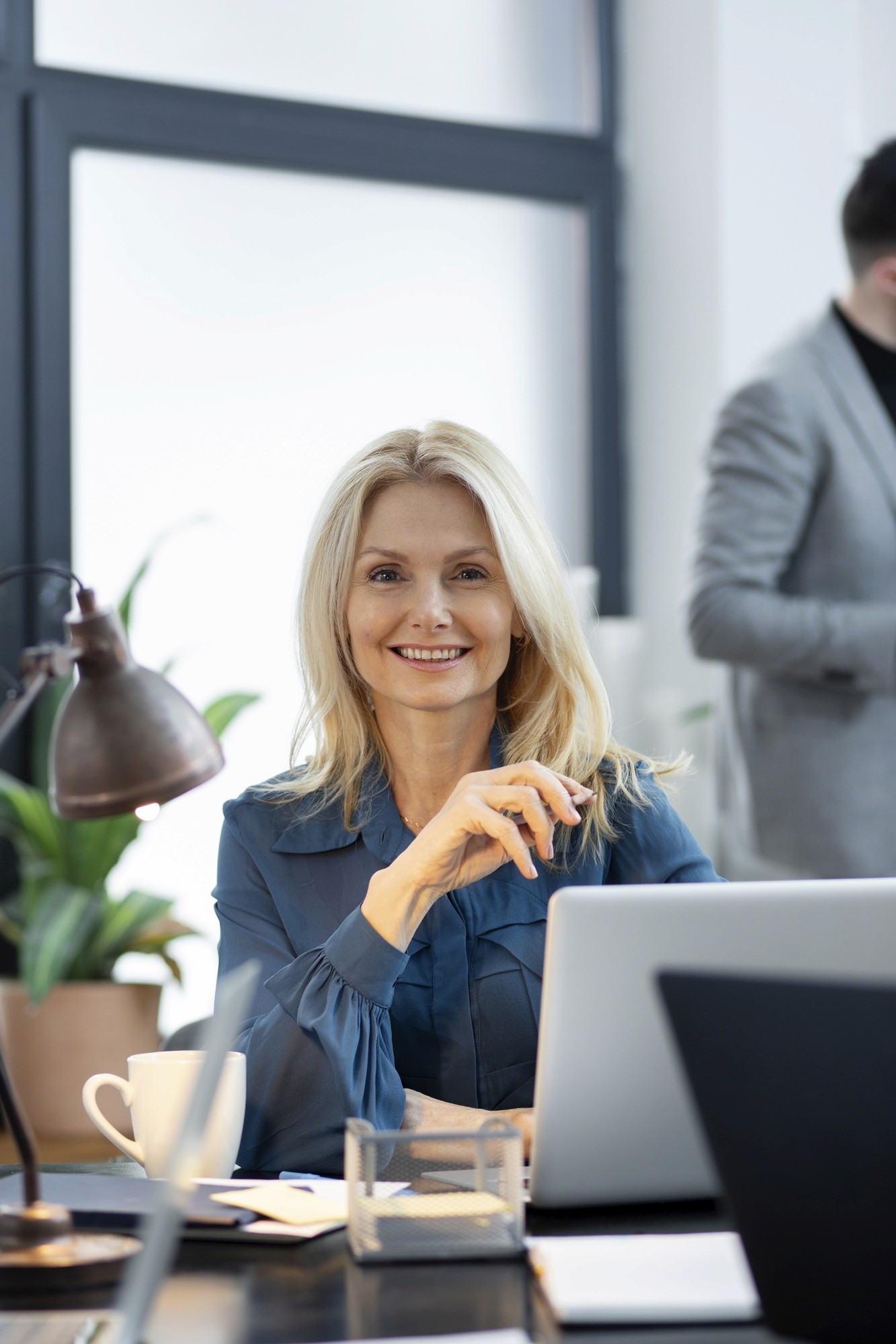 A smiling blonde woman sitting at a desk with a laptop, a cup, and office supplies, in a modern workspace