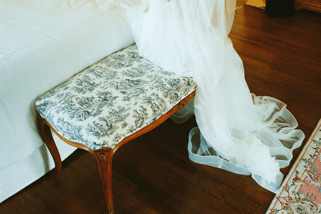 Bridal veil and gown draped beside a wooden bench with a green toile cushion on a dark wood floor.