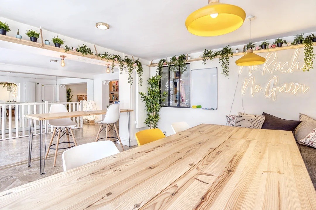 Espace de coworking Alveus avec une grande table commune en bois massif, luminaires suspendus jaunes et décoration végétale.