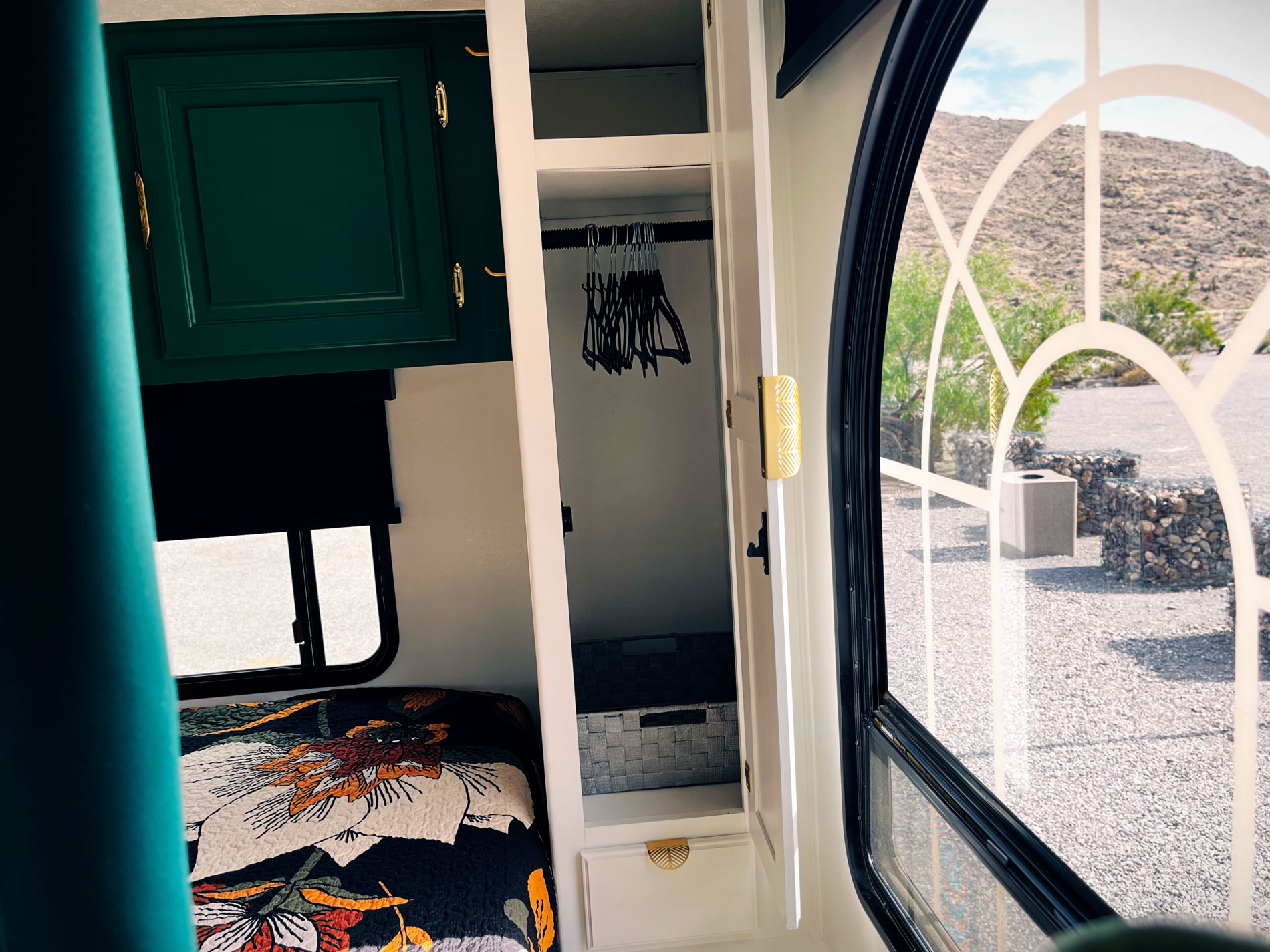 Cozy RV interior with a floral bedspread, green cabinet, and open closet with hangers. Sunlight filters through an arched window, framing a desert view.