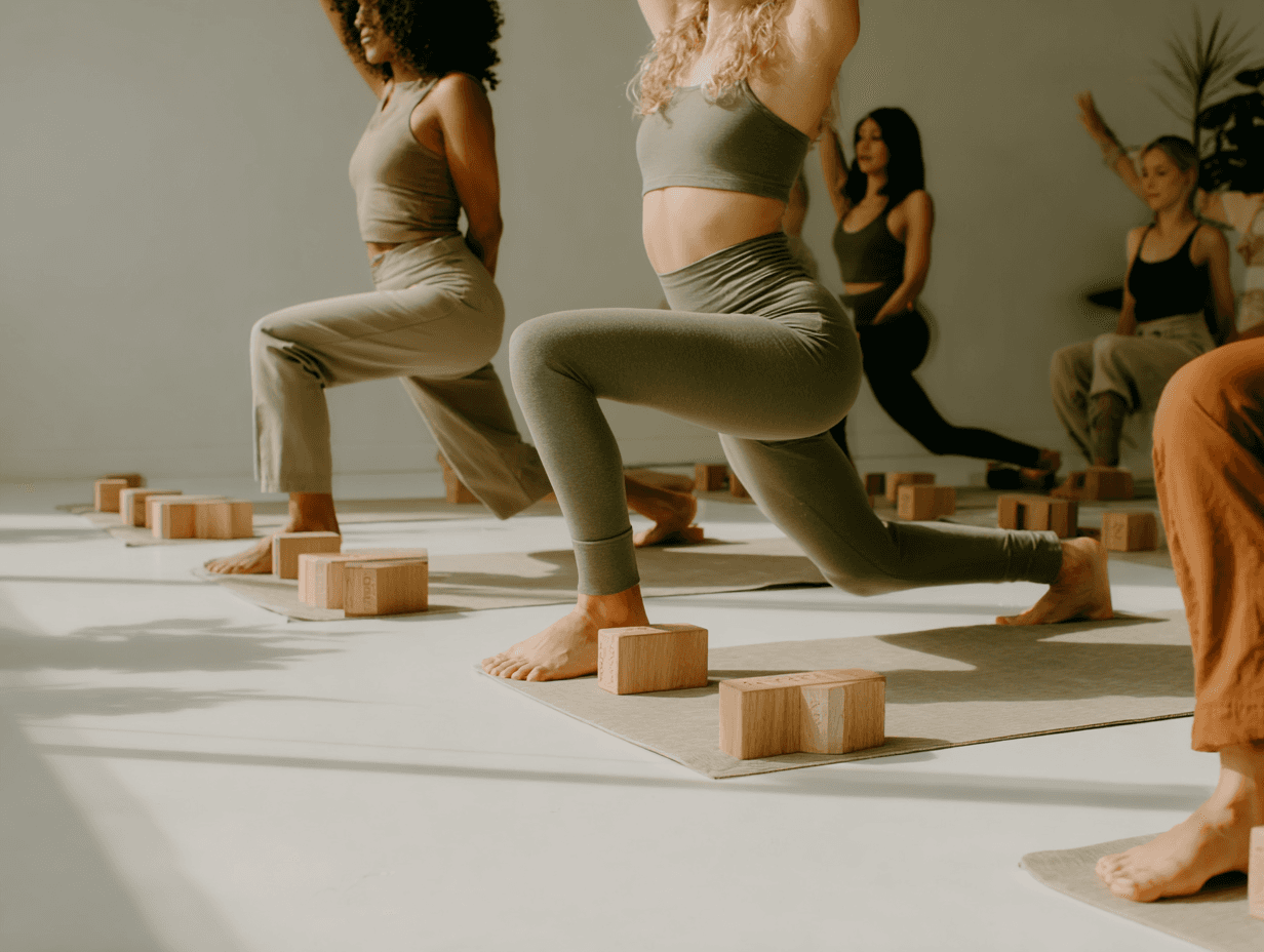 A group of women in coordinated yoga poses during a sunlit class, with wooden yoga blocks and soft-toned outfits in a minimalist studio