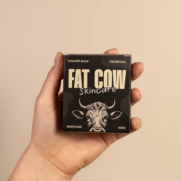 A hand holding a black soap box labeled 'FAT COW Skincare' featuring an illustrated cow's face. The box indicates it's a 100g tallow soap with charcoal.