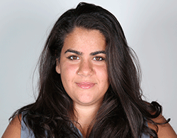 A woman with long, dark brown hair and brown eyes looks directly at the camera. She has a neutral expression and is wearing a light blue top against a plain, light gray background.