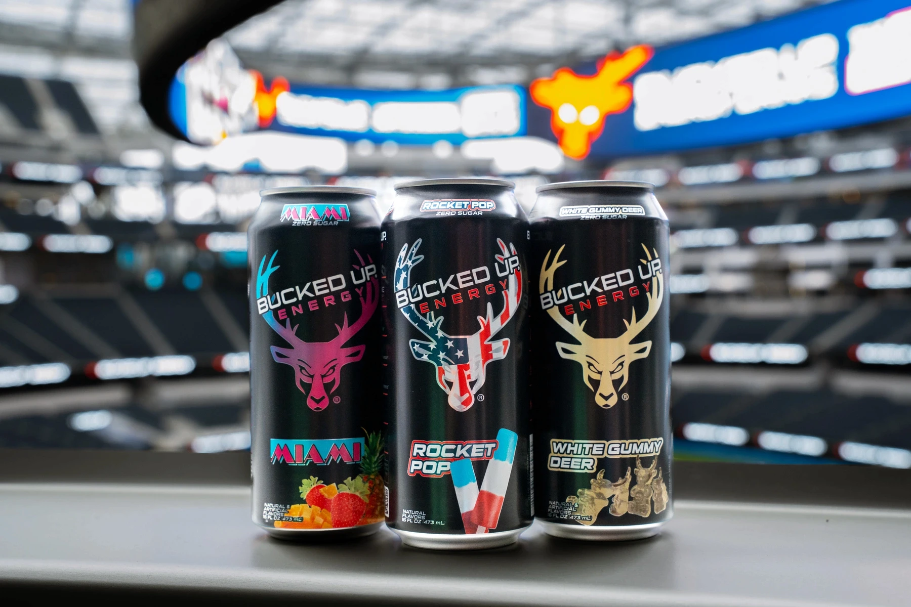 Three Bucked Up Energy drink cans lined up in front of a stadium background featuring flavors Miami Rocket Pop and White Gummy Deer with signature deer head logo.