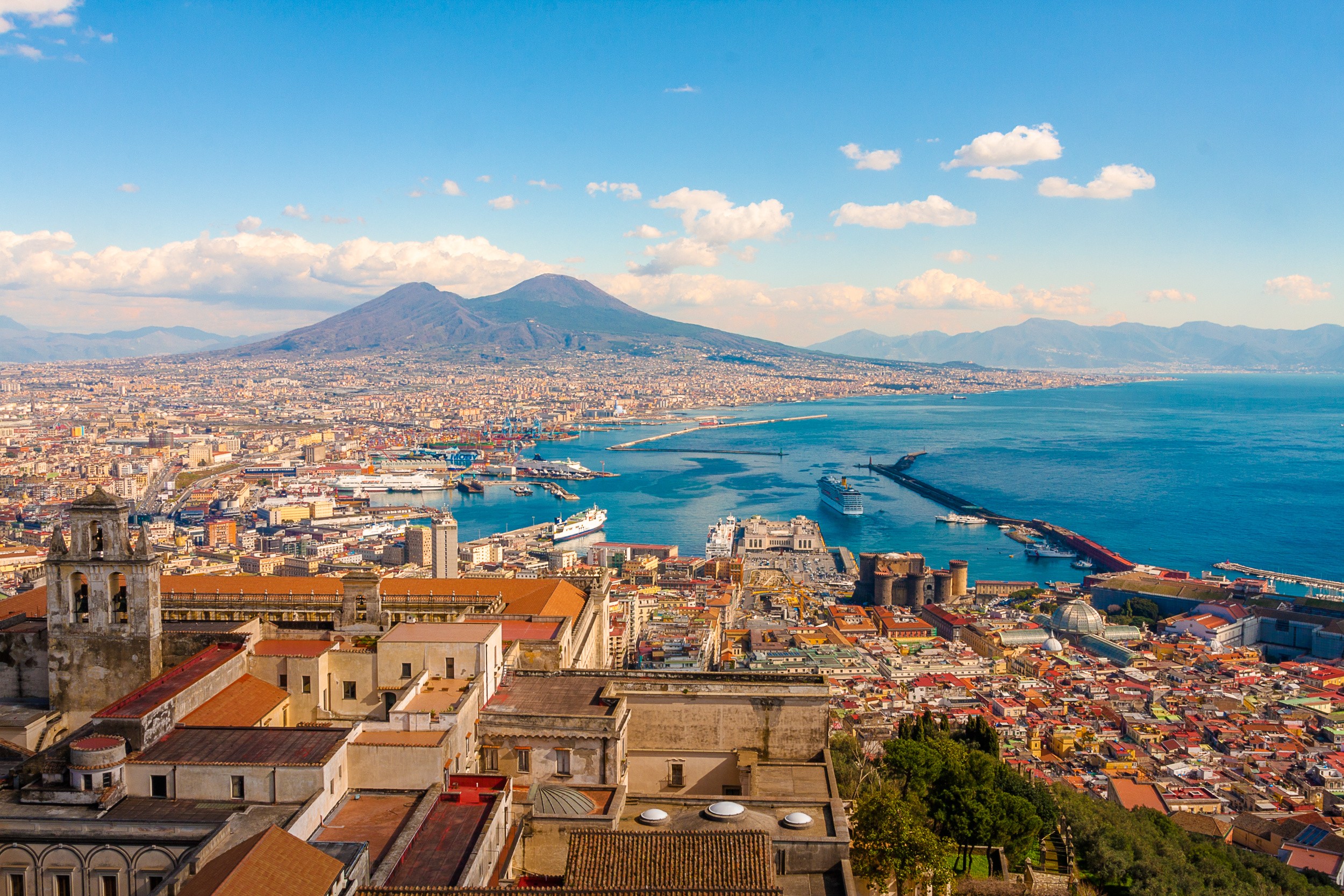 Vista panoramica di Napoli, Italia, con il paesaggio urbano storico, il porto animato e l'iconico Vesuvio sotto un cielo azzurro con nuvole sparse.
