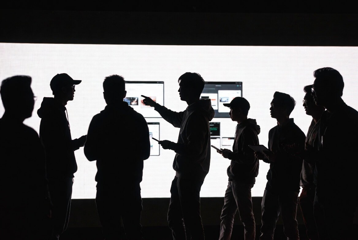 Creative team in deep refinement session. Silhouetted group closely examining UI interface details on a large screen, with team members pointing and gesturing to polish every pixel and interaction. Dramatic backlit black and white photo.