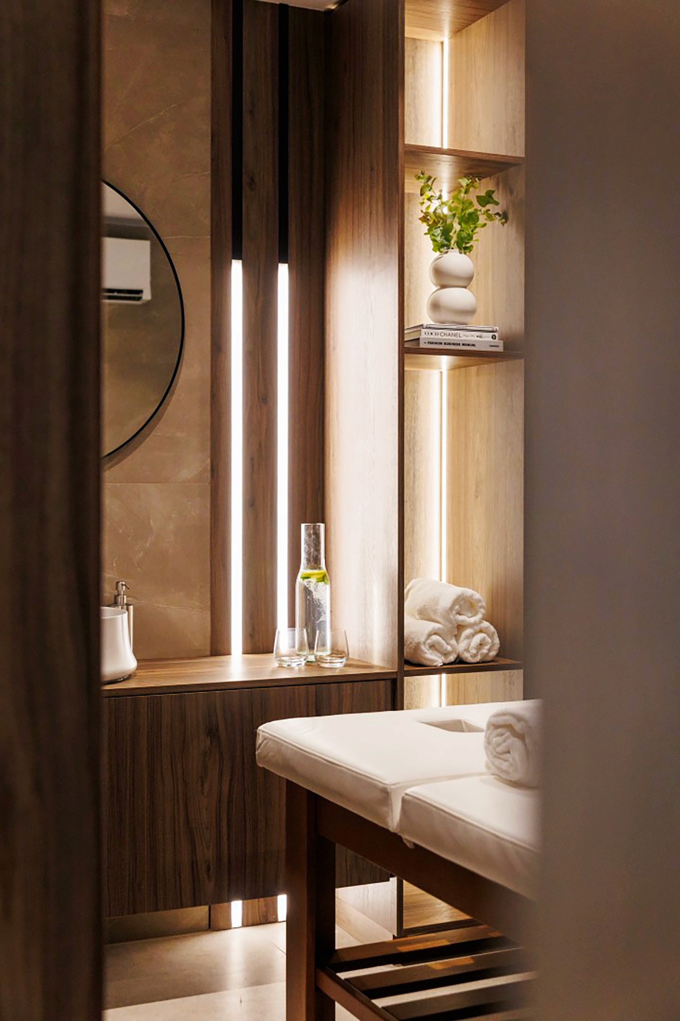 Villa Trio spa room detail with massage bed, warm wood finishes, and softly lit shelving