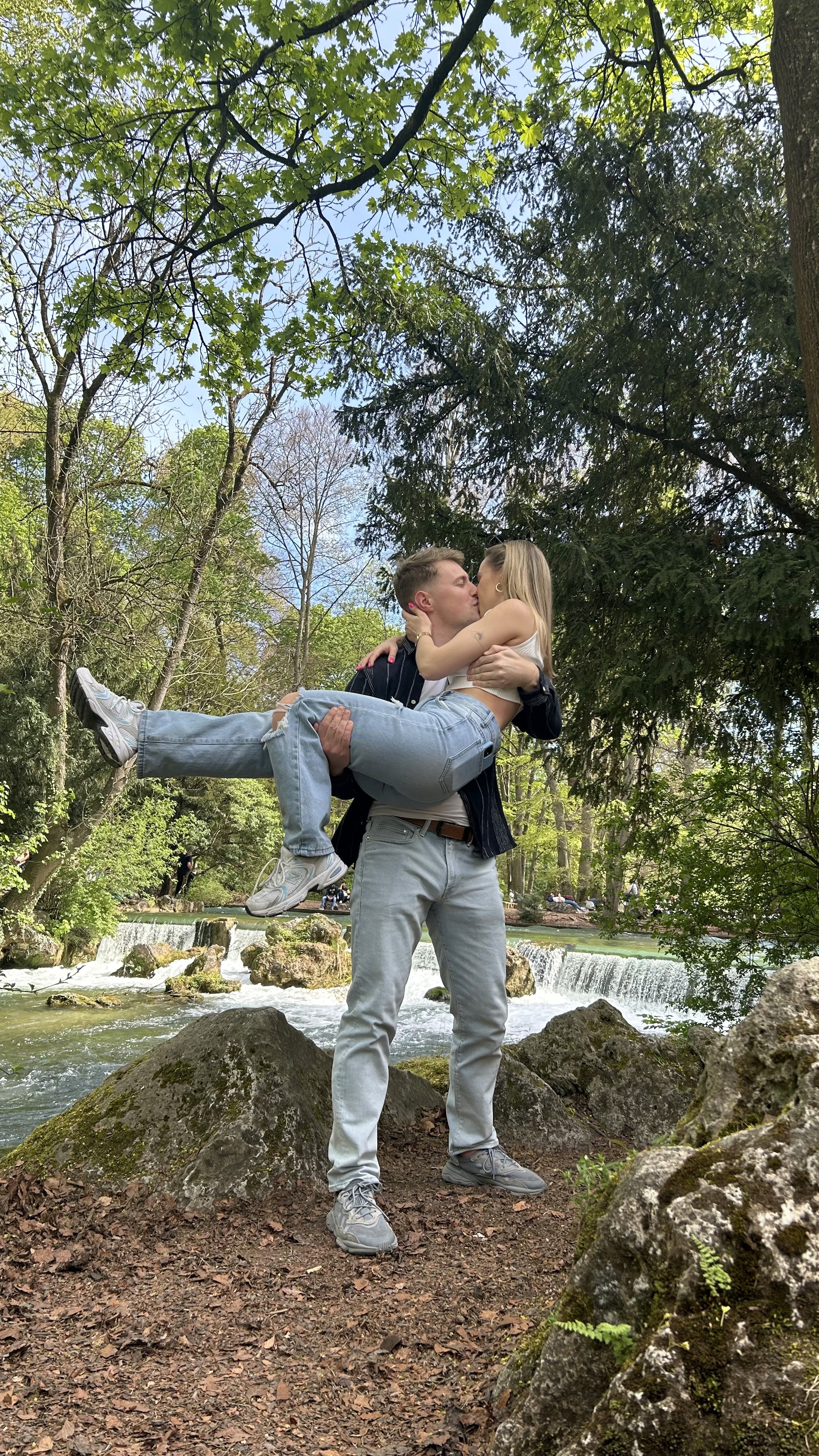 A groom in a dark suit carries a bride in a white dress across a sunny, grassy field. The couple shares a joyful, intimate moment with trees in the background.