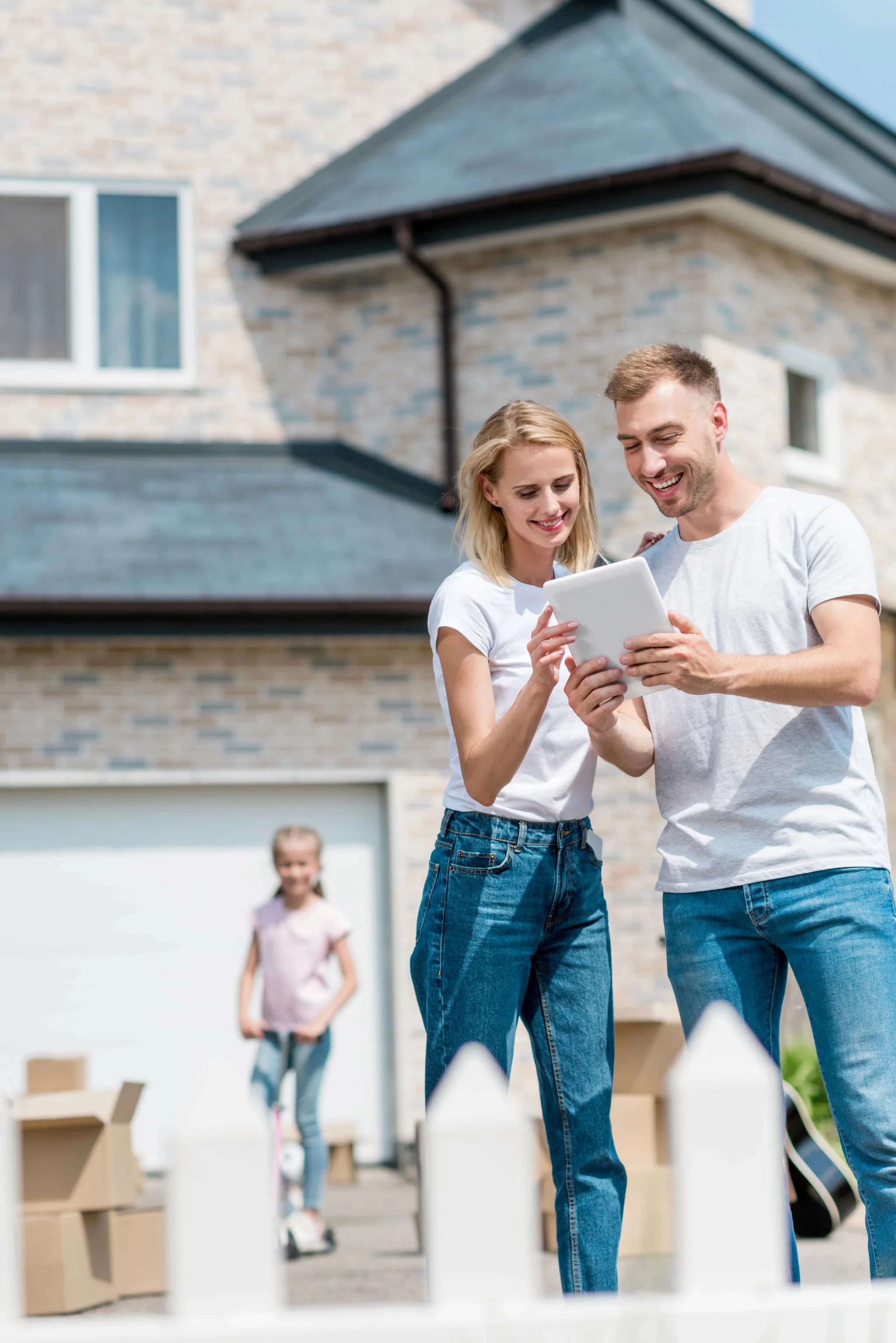 A young couple in casual clothing smiles while viewing a tablet outside their new brick home, with a child and moving boxes visible in the background.