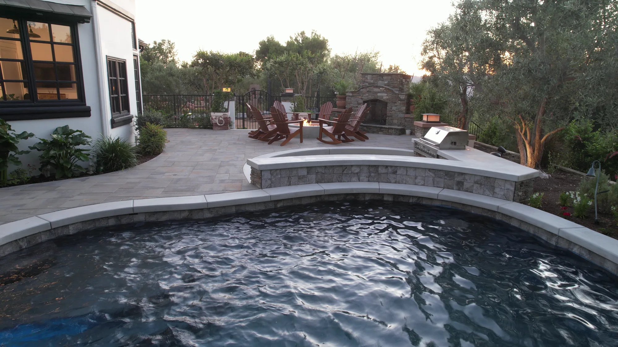 Curved swimming pool in the foreground with a paved seating terrace and stone fireplace beyond.