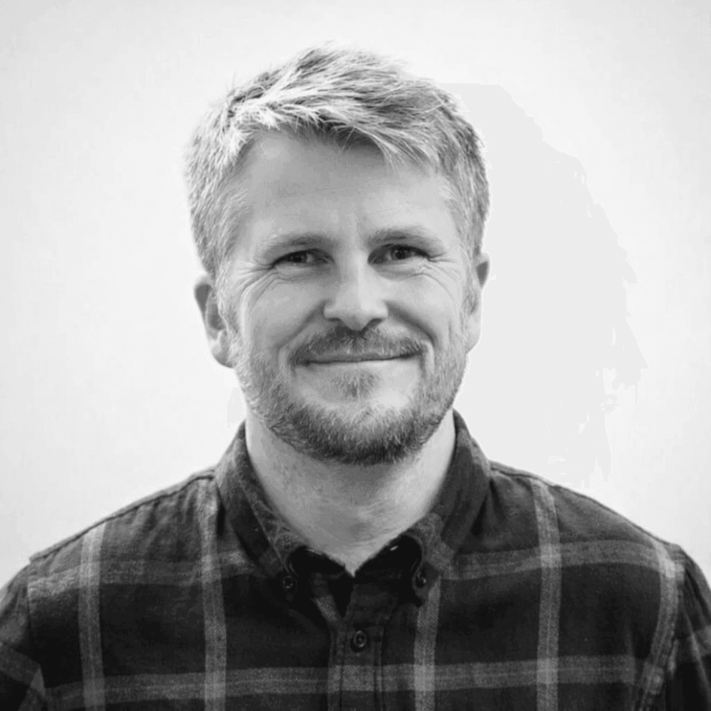 Portrait of a young man in a dark double-breasted suit and black turtleneck. Soft lighting creates a sophisticated, serious tone.