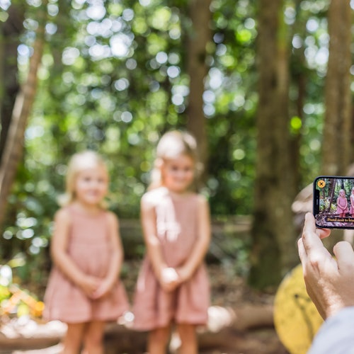 Dua gadis muda dalam gaun merah muda yang serasi sedang difoto di lingkungan hutan. Fokusnya adalah pada ponsel pintar yang mengambil foto tersebut.