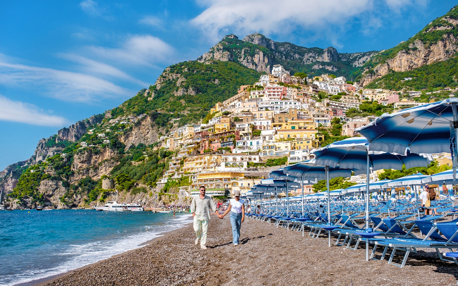 Couple walking on Positano beach with colorful hillside buildings, Amalfi Coast.