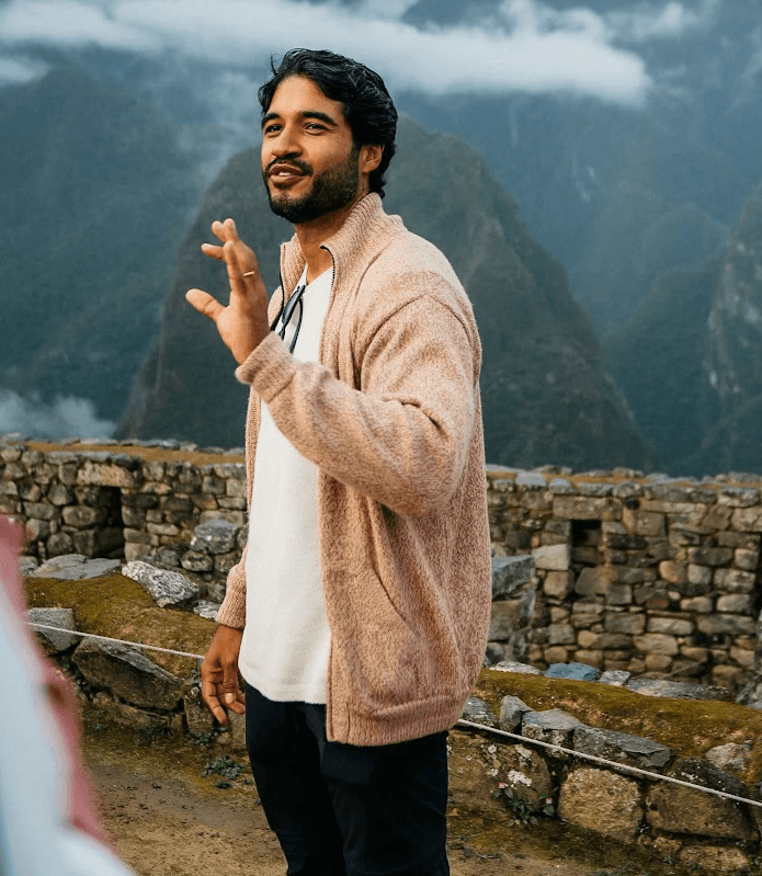 A smiling young man with crossed arms, wearing a plaid shirt and white t-shirt, poses against a dark background.