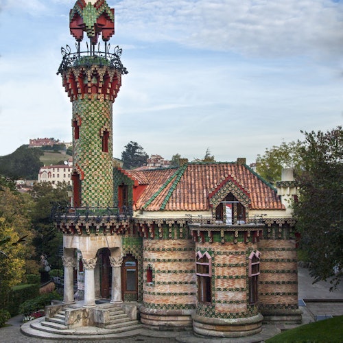 Un edificio decorativo con un techo de tejas rojas y una torre cubierta de mosaicos, rodeado de vegetación en un día soleado.