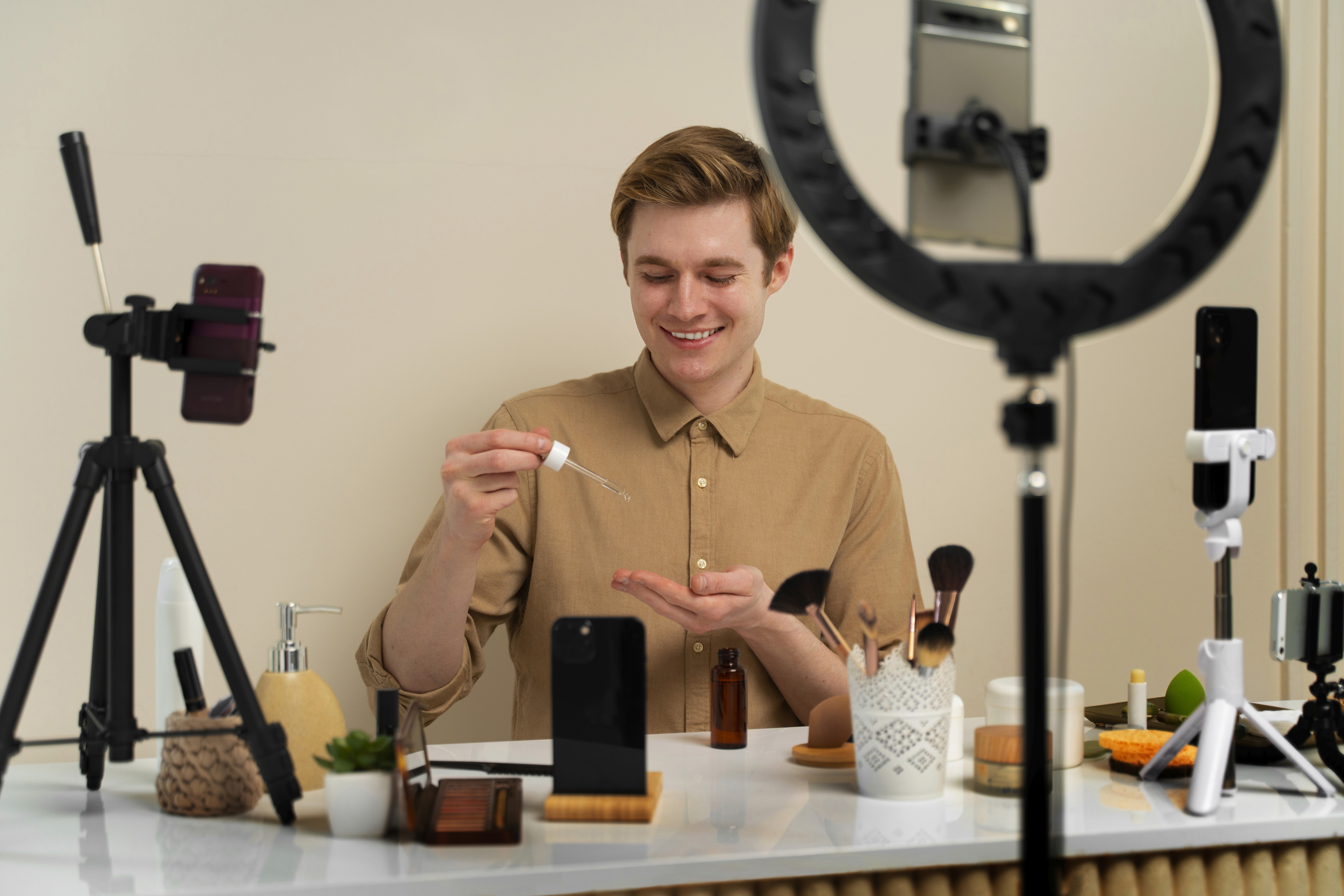 A male influencer smiles while demonstrating skincare serum at a desk with multiple smartphones, tripods, and a ring light
