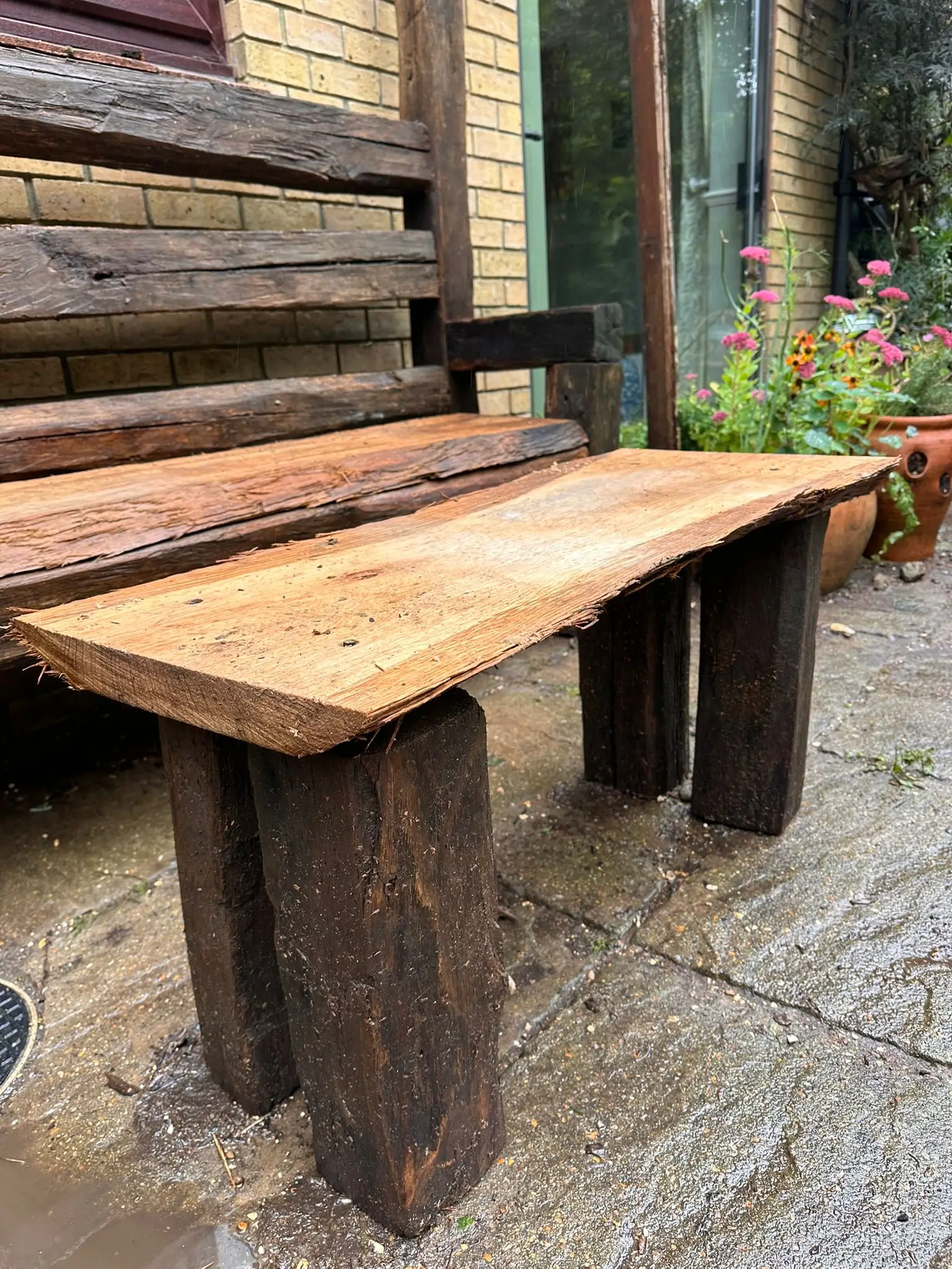 A rustic wooden bench with a flat surface, supported by sturdy log legs, set outdoors against a natural backdrop.
