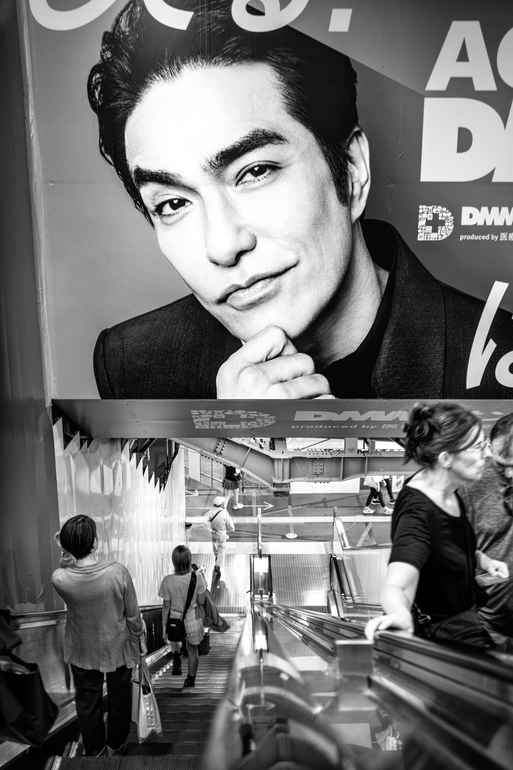 Black and white photograph of people riding an escalator beneath a large advertisement in a department store.
