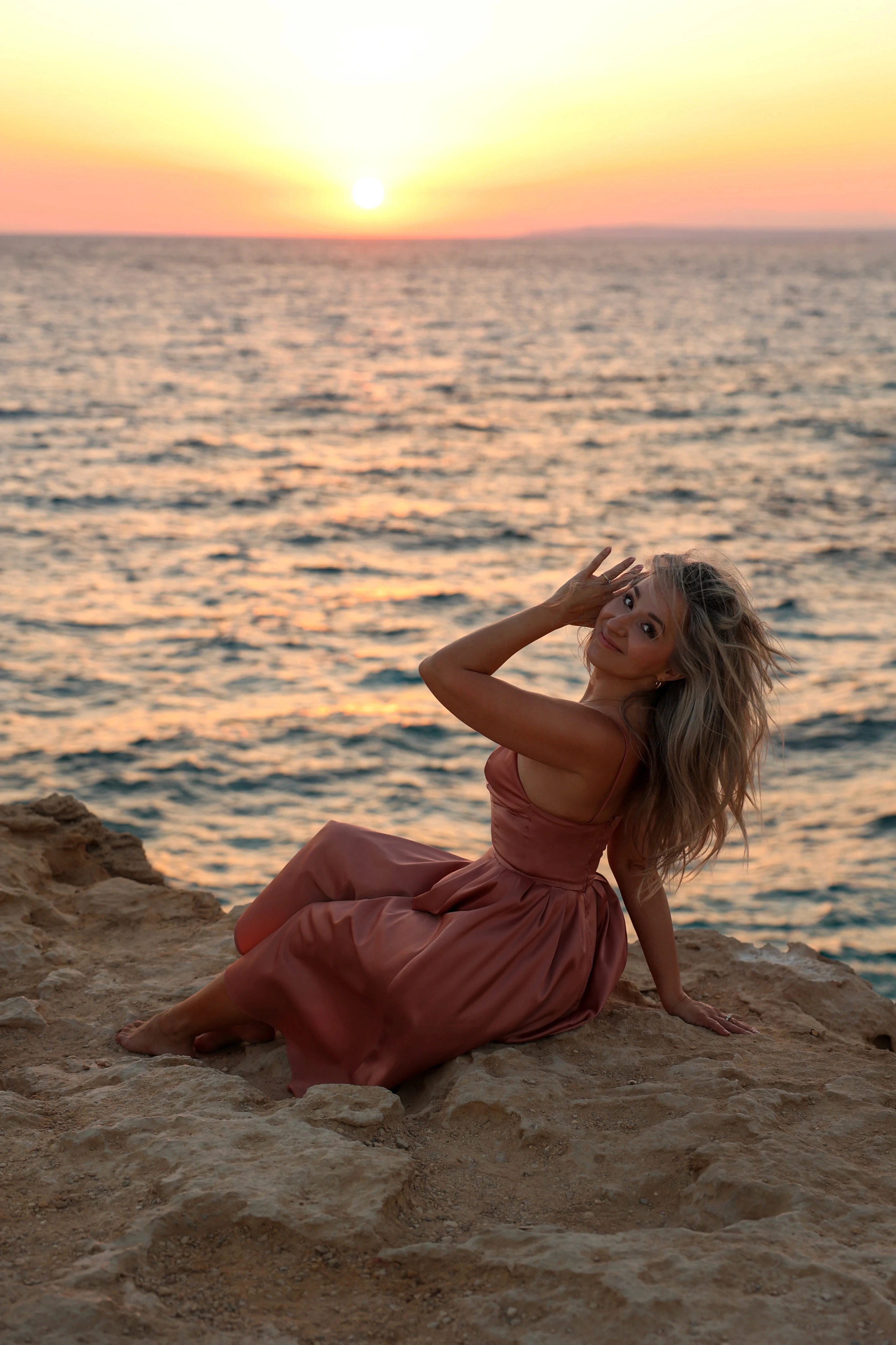 Radiant woman in a rose gold silk dress sitting on rocky cliffs, embracing her inner light during a golden hour sunset by the sea.