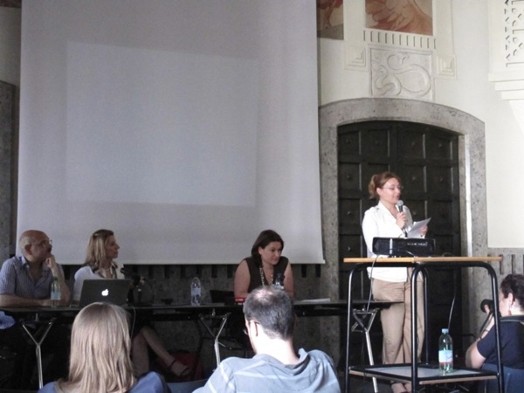 A photo of a woman giving a lecture while reading something of a piece of paper.