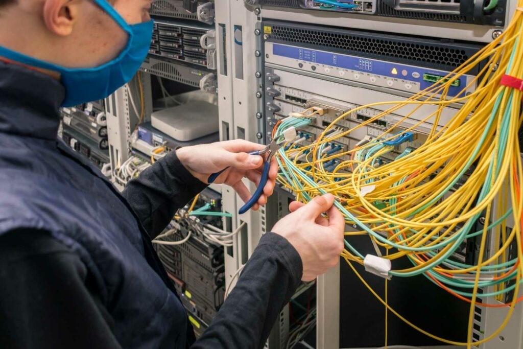 Técnico de redes com máscara manuseando cabos de fibra óptica em rack de servidores, representando instalação e manutenção de infraestrutura de rede em data center - FindUP -Manutenção preventiva e corretiva
