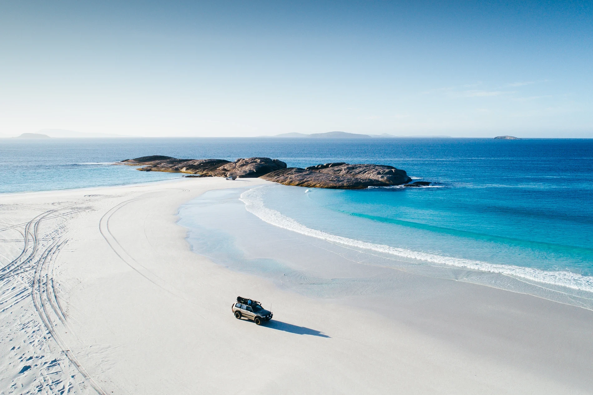 A four-wheel drive on a white beach beside turquoise water and granite rocks