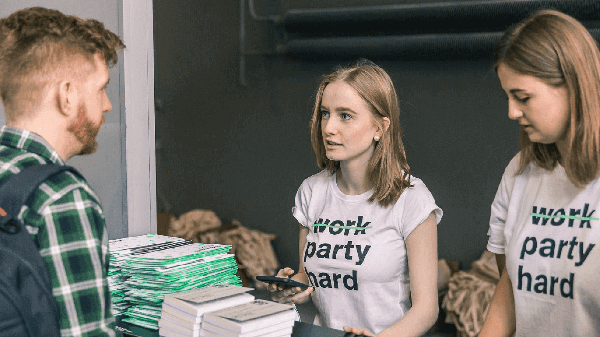 Mężczyzna rozmawia z dwiema kobietami przy stosie zielono-białych materiałów; na ich koszulkach „work” jest przekreślone nad „party hard”.