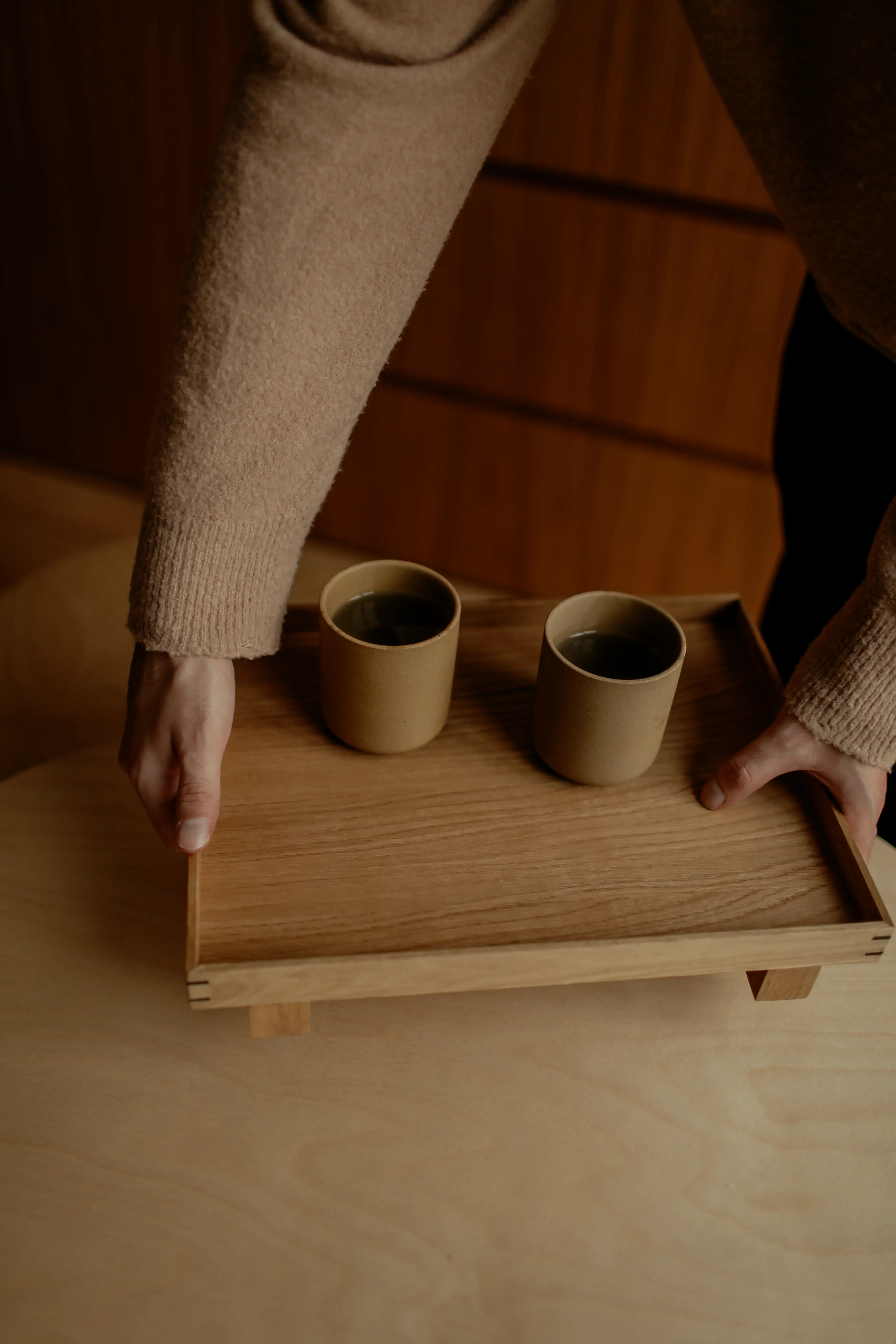A person holding a wooden tray with two ceramic cups on it