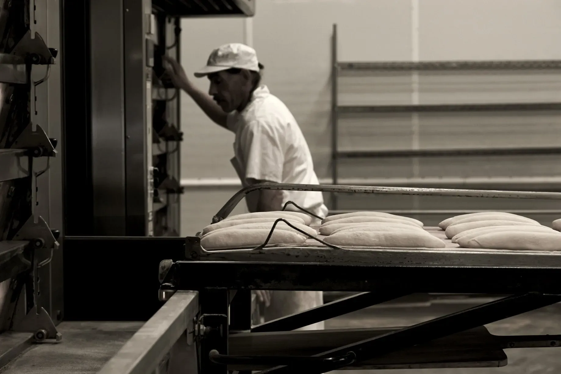 Fresh baguette flour about to enter the oven, baker prepares oven in background