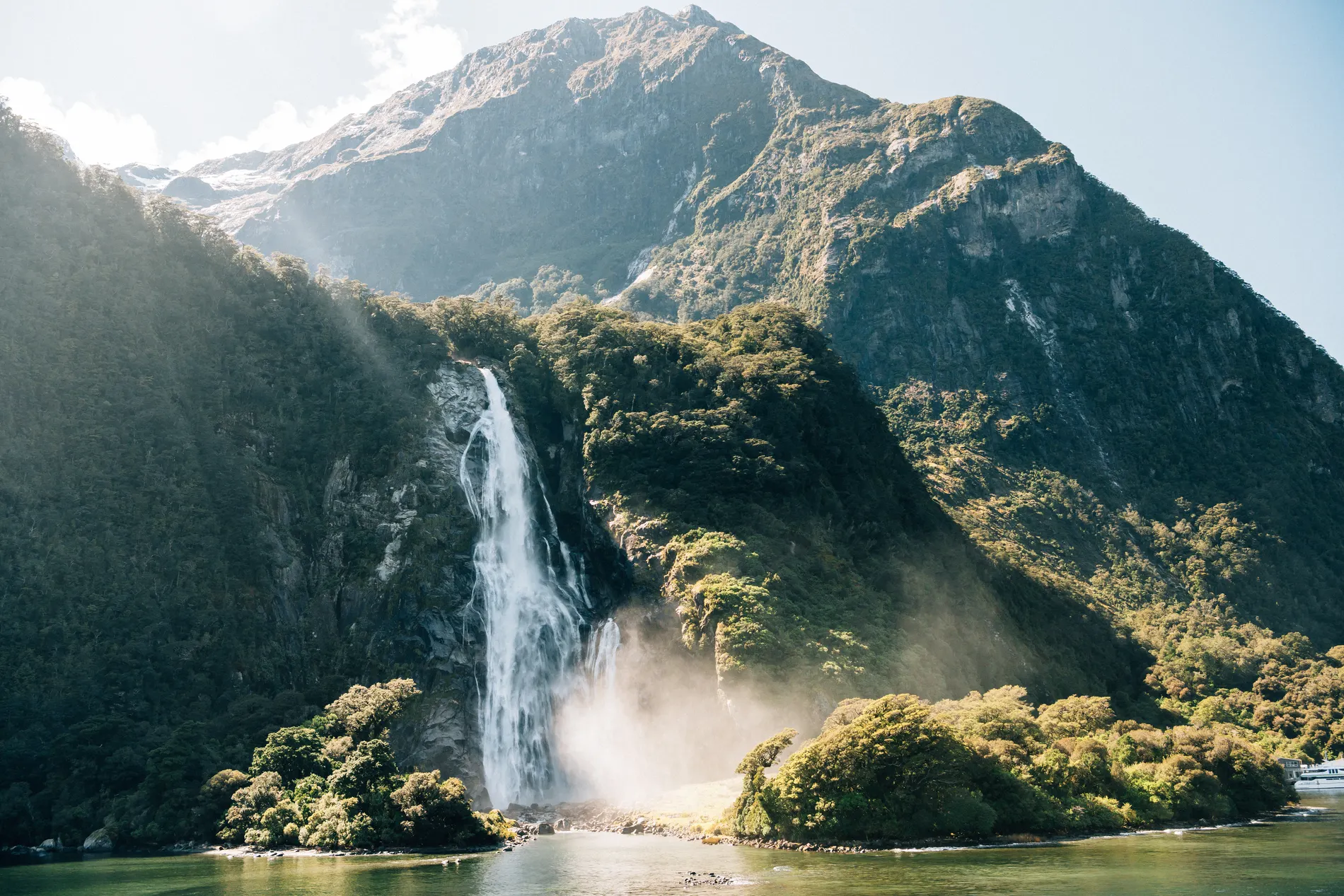 A tall waterfall falling into a forested basin, sunlight breaking over the ridge