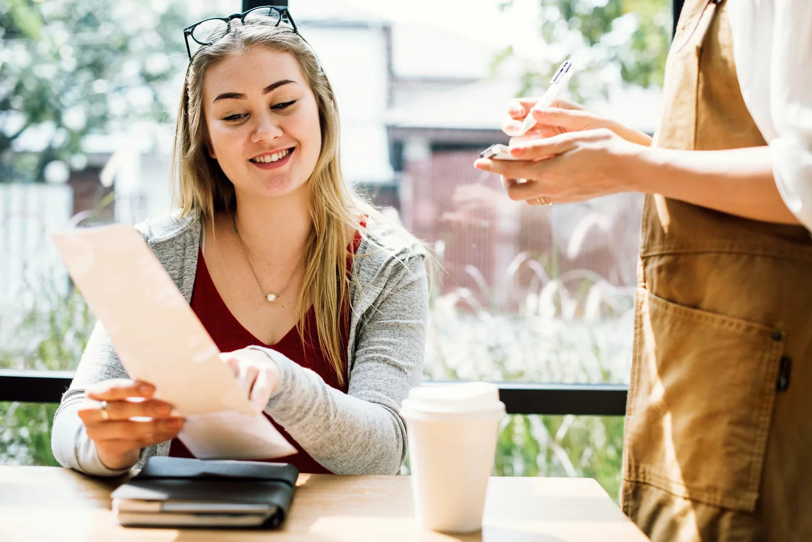 Young woman smiling while reviewing documents at a café table with a colleague holding a brown paper bag.