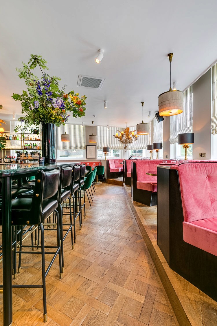 A chic restaurant with pink velvet booths, green barstools, and vibrant floral arrangements.