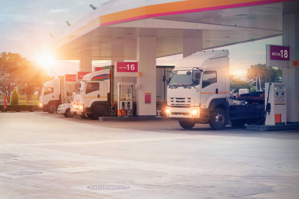 Fleet of tank trucks at a fueling station with fluctuating fuel price signage in the background.  