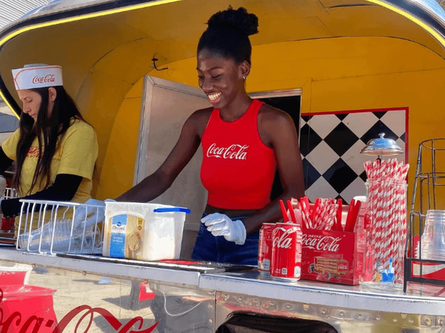 Two women work at a retro-style food truck. One wears a red Coca-Cola tank top and gloves, smiling as she prepares drinks with straws and napkins on the counter. The other wears a yellow shirt and a white hat.