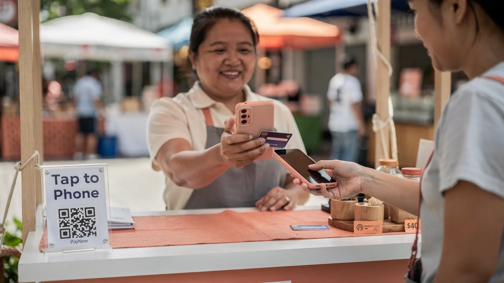 A Singapore small business owner at a pop-up booth holding an Android phone with an NFC contactless payment screen, while a customer taps a credit card on the back of the phone. A simple “Tap to Phone” sign is visible next to a PayNow QR code on the counter.
