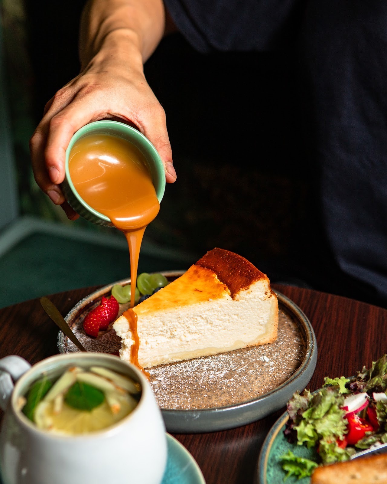 Eine Hand hält ein kleines Kännchen und gießt eine reichhaltige Karamellsauce über ein Stück Käsekuchen auf einem dunklen Teller, garniert mit frischen Früchten.