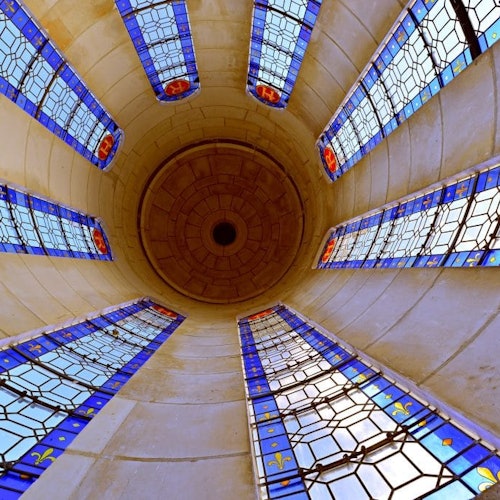 Un plafond voûté et orné avec des sculptures minutieuses, trois grandes fenêtres cintrées, et la lumière du soleil inondant une salle vide au sol en pierre.