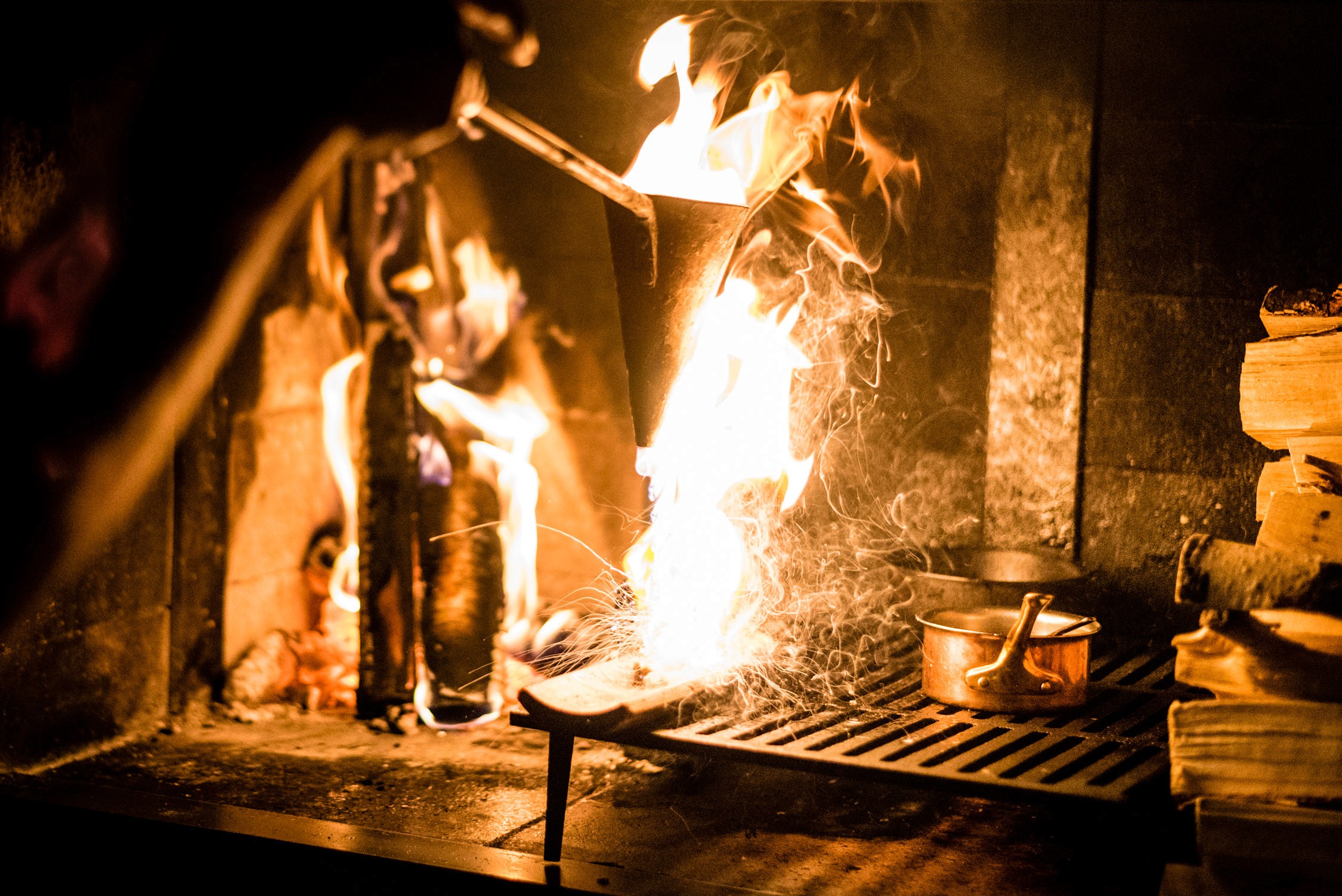A person in a dark apron stirs a pot over an open flame on a metal grill inside a rustic stone kitchen.