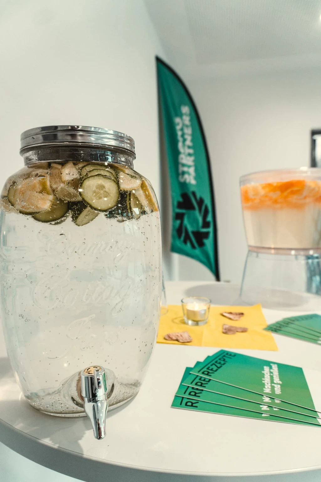 Eine großer Wasserspender mit Gurken drin steht auf einem Tisch, auf dem auch Strong Partners Flyer liegen