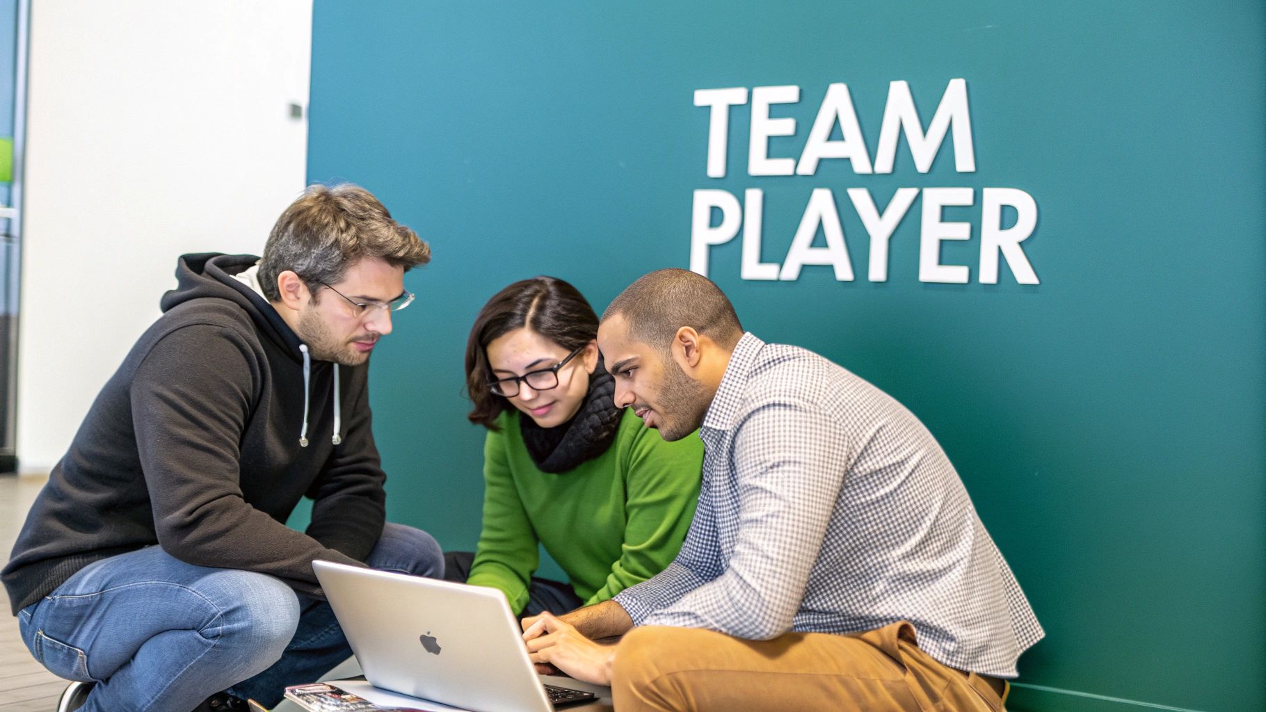 Three young professionals, two men and one woman, collaborating around a laptop on the floor, in front of a 'TEAM PLAYER' wall.