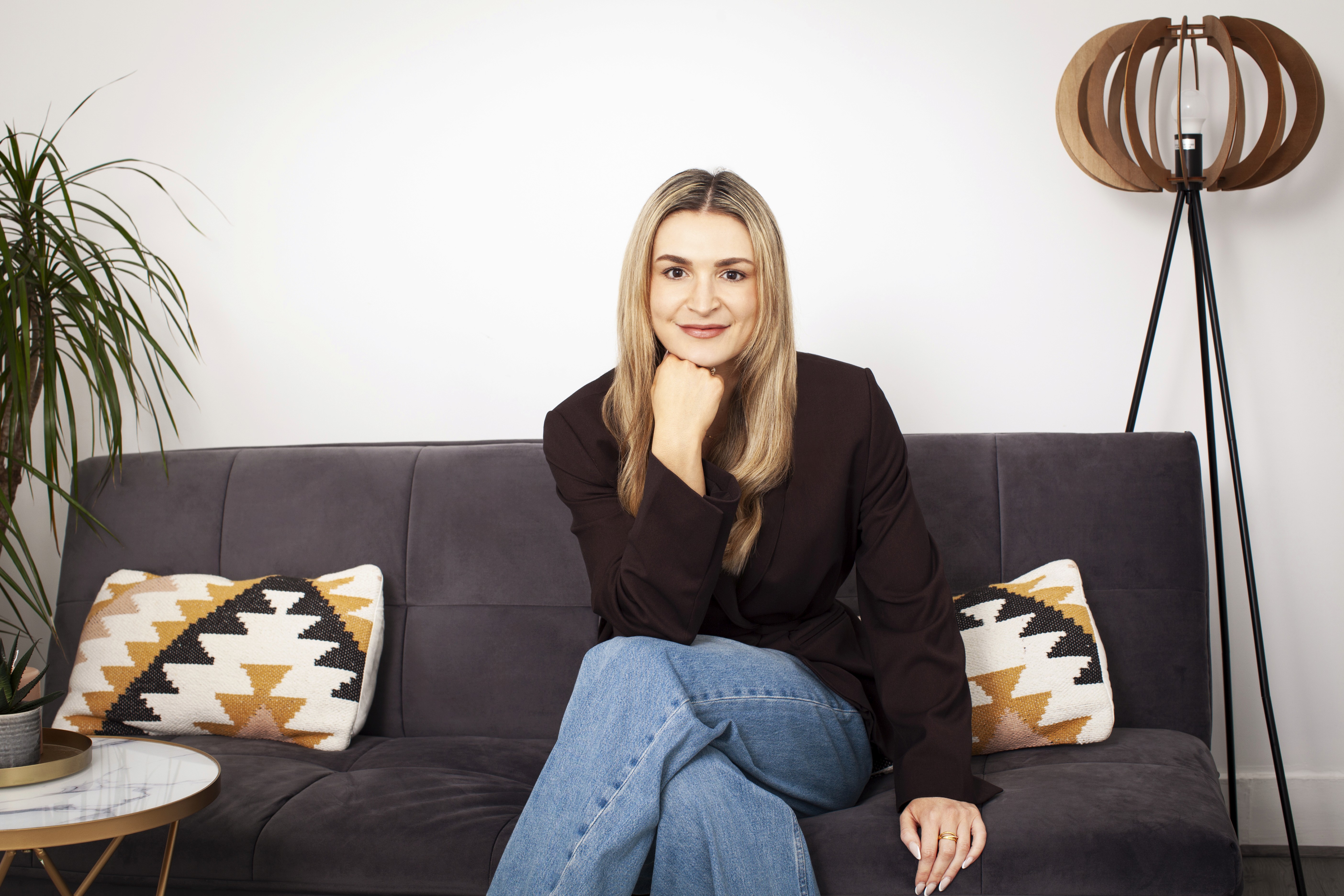 Woman sitting on a sofa with her chin resting on her hand, smiling at the camera, with a plant and floor lamp in a modern living room.