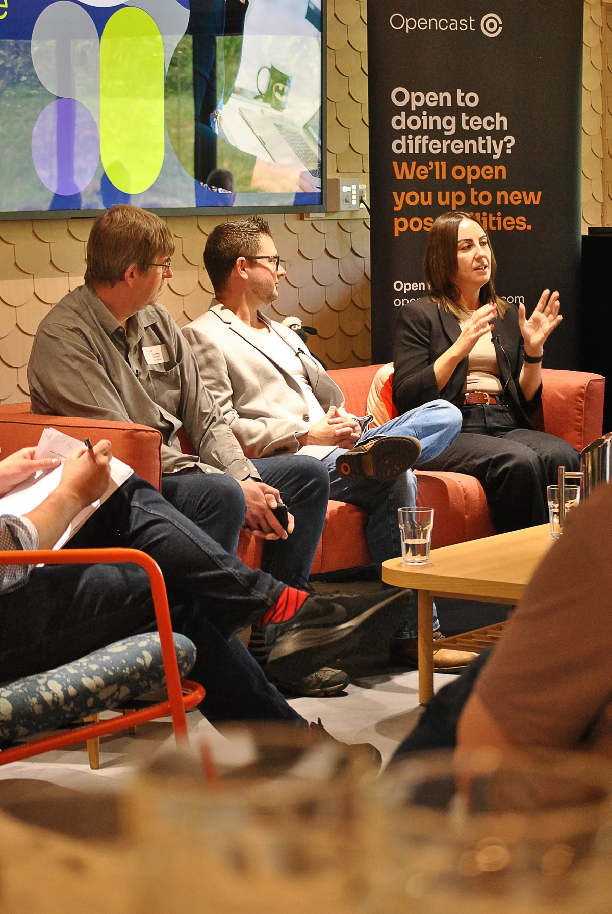 Three people seated on a sofa during a tech event, with an Opencast banner and a large screen in the background displaying graphics and text.