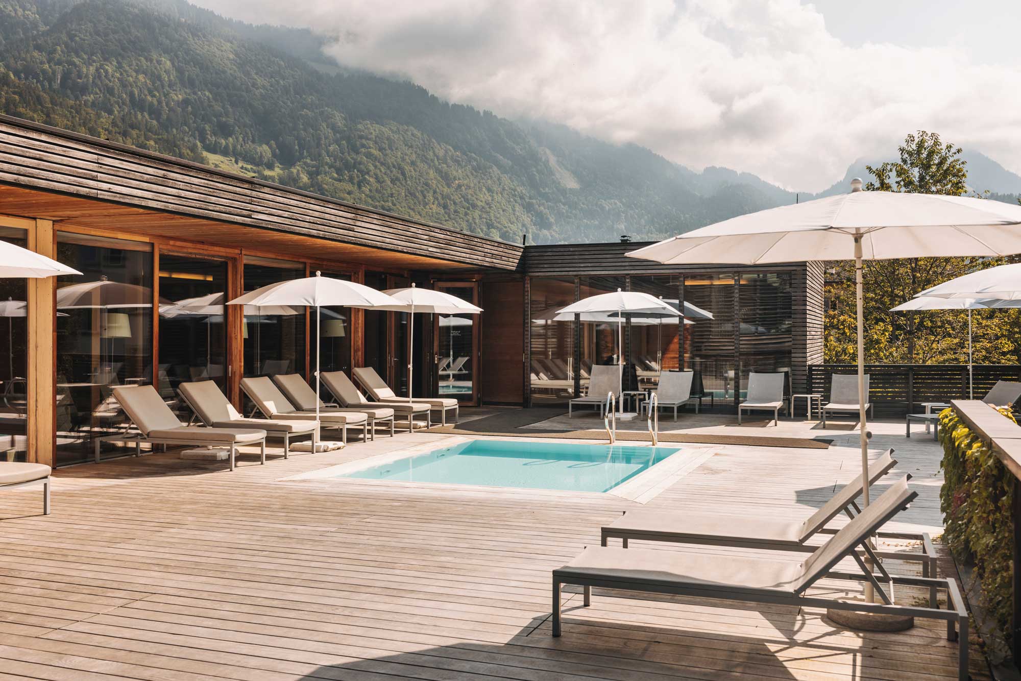 Expansive spa terrace with rectangular pool, multiple sun loungers, white umbrellas, and dramatic Alpine mountain backdrop.