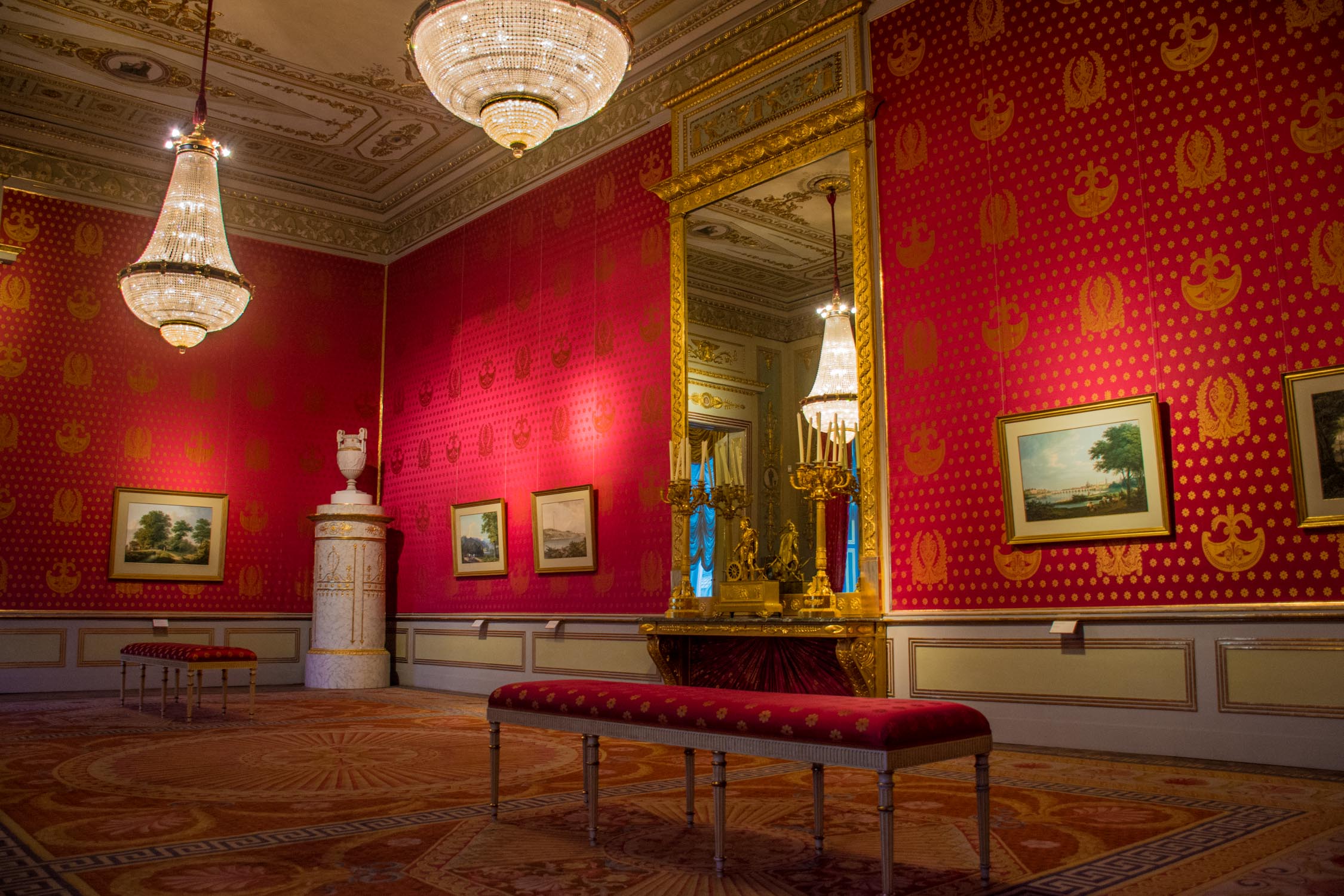 A red and gold patterned wallpaper room features a long velvet sofa, ornate chandeliers, and framed paintings.